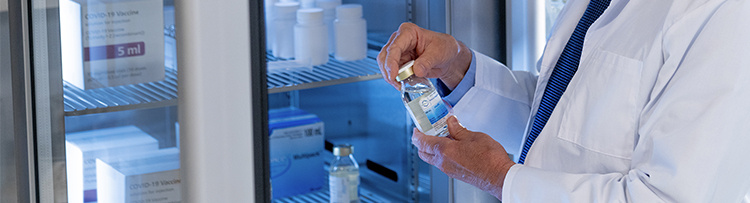 Vaccine close up of vial in doctor's hand standing in front of refrigerator with more vaccines